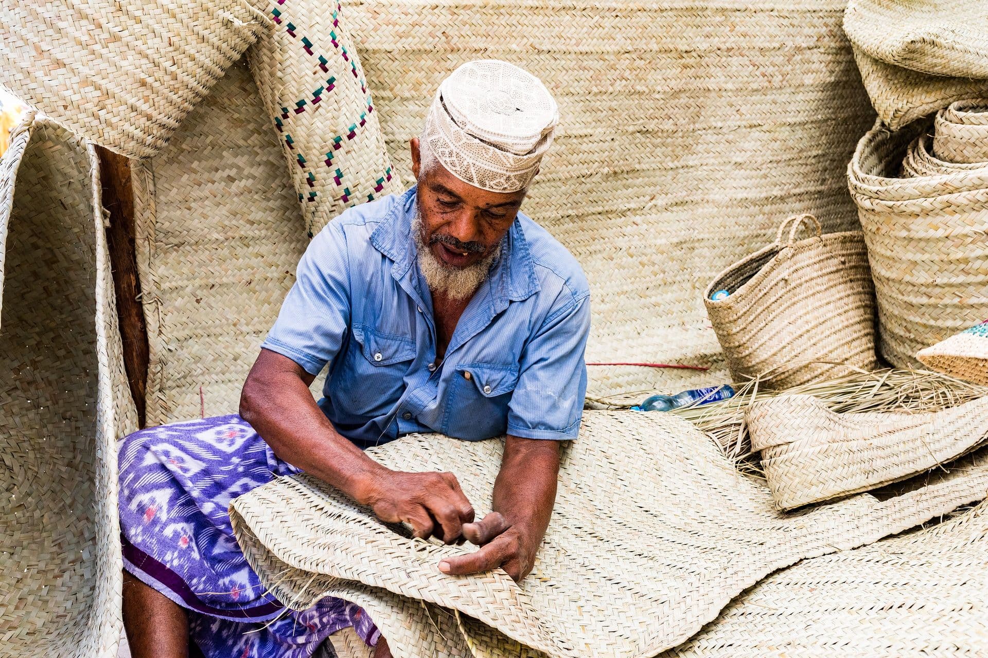 An elderly man in a white kofia weaving a palm-leaf mat, surrounded by woven baskets