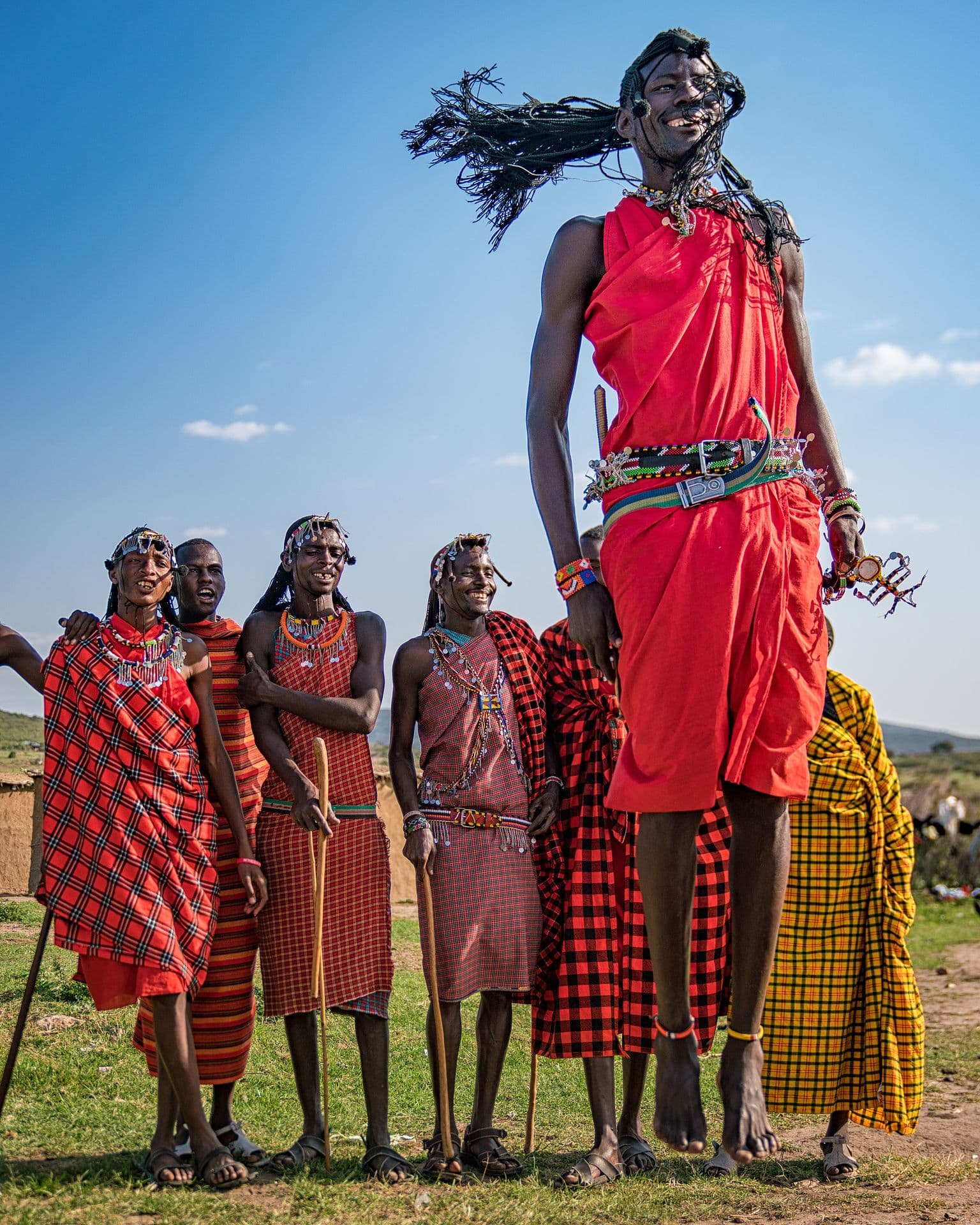 A Maasai man in red shúkà leaping high above a group of men watching on a grassy hill
