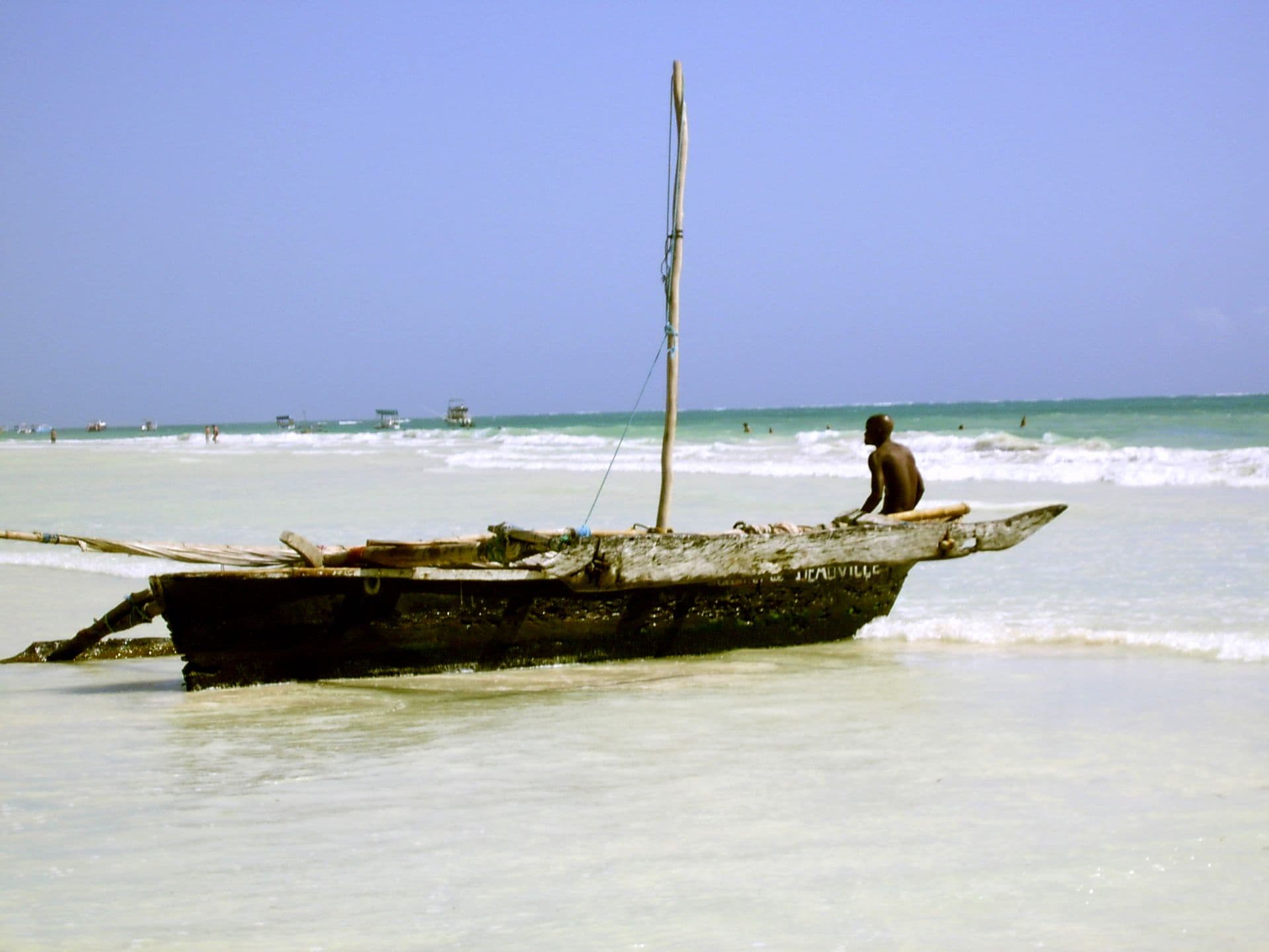 A wooden outrigger canoe beached in shallow water at Diani, with a man standing beside the mast