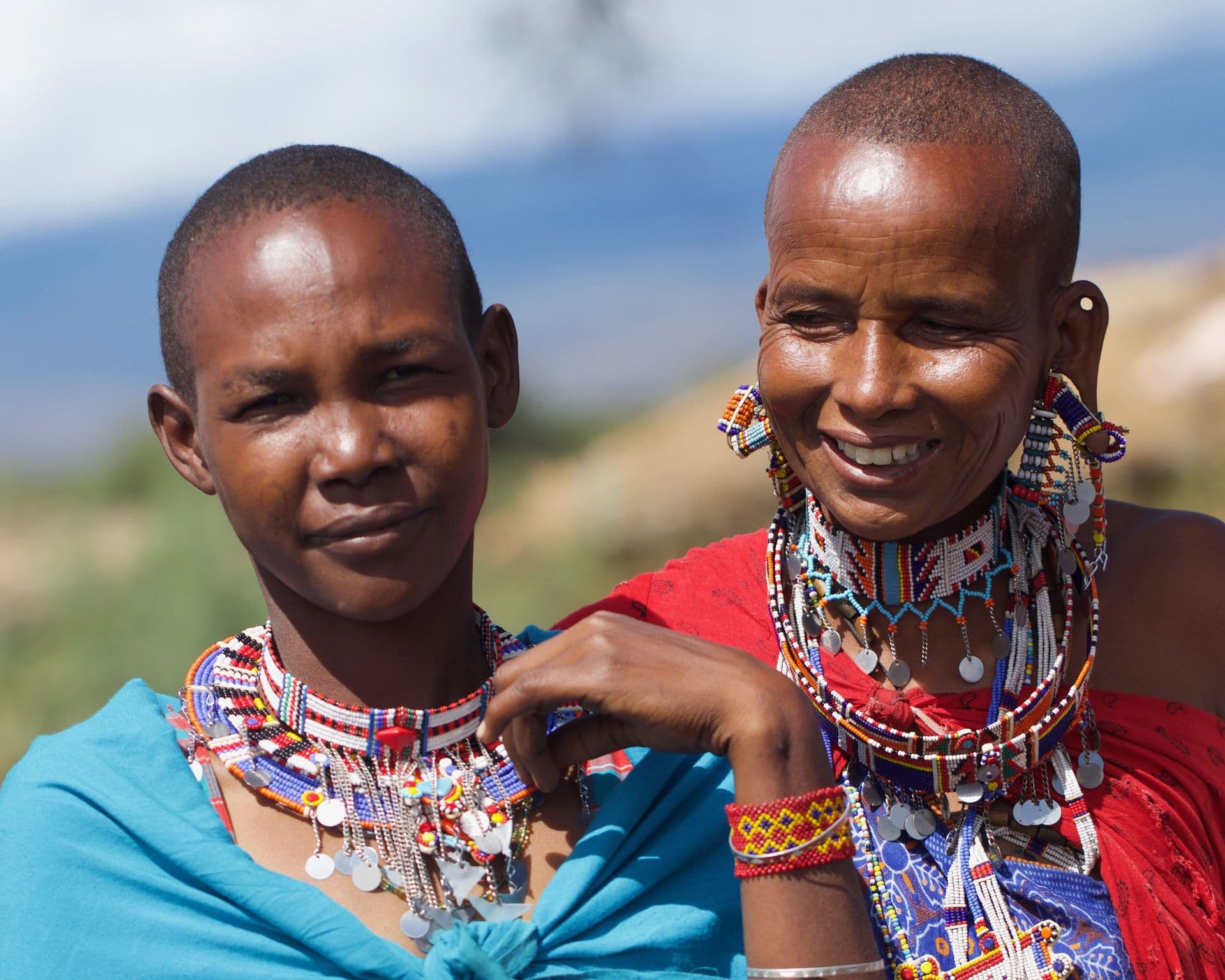 Two smiling Maasai women wearing wide beaded collars and blue and red shúkà