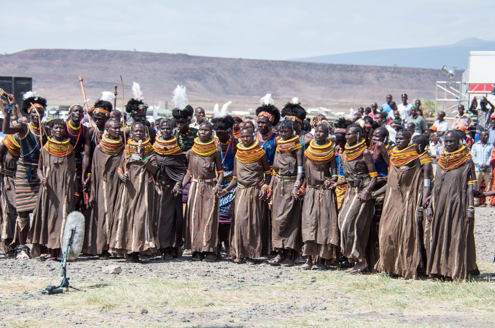 A long row of Turkana women in beaded collars and brown wraps dancing on open ground